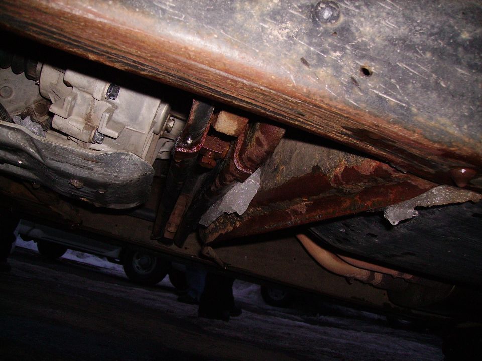 Underside of a vehicle showing rust and a transmission. Another vehicle is visible in the background
