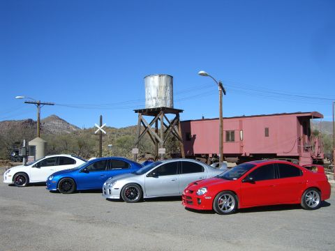 1996 Ford Probe - Four Dodge Neon SRT-4s lined up in front of a train car and water tower under a clear blue sky.