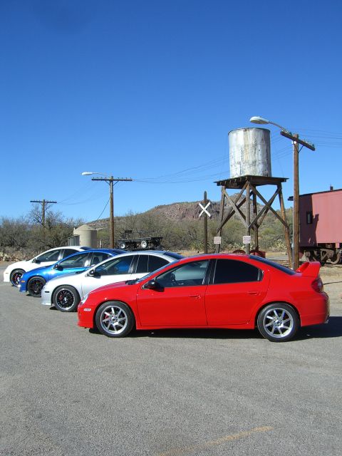 1996 Ford Probe - Several sedans are parked in a line on a paved lot with a water tower and train car in the background.