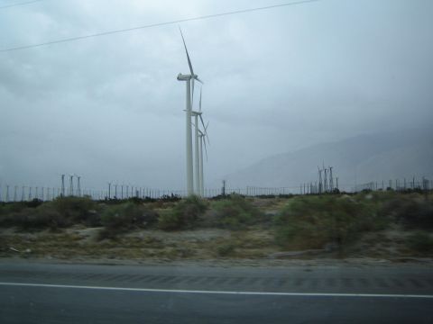 1990 Ford Bronco - Tall wind turbines rise from desert scrub beside a roadway, with hazy mountains behind under a gray sky.