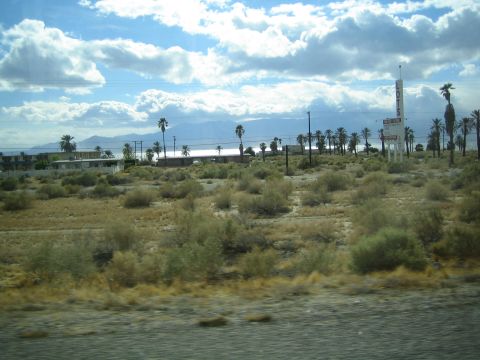 1990 Ford Bronco - desert roadside scene with palm trees, low buildings and a sign overlooking a distant lake under puffy clouds.