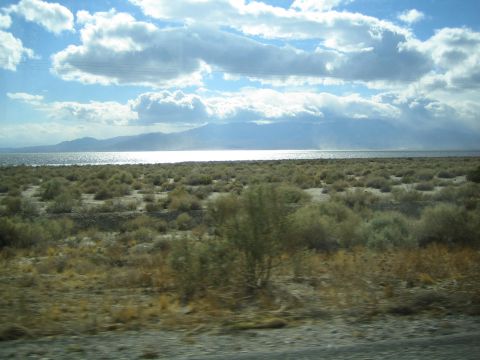 1990 Ford Bronco - sunlit desert lake stretches toward distant mountains under scattered clouds, framed by roadside brush.