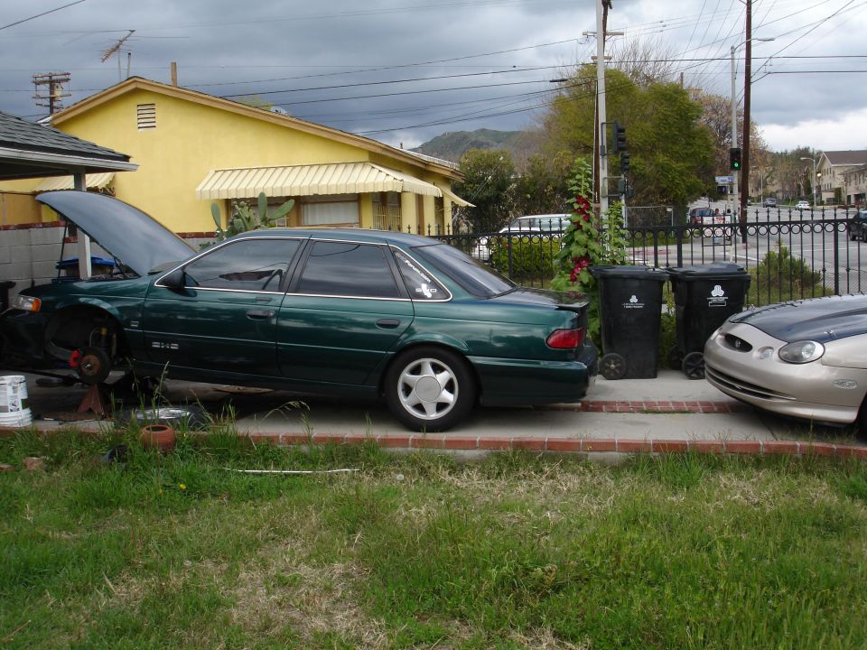 Green 1993 Ford Taurus sedan with hood open, undergoing repairs. Another car is parked to the right