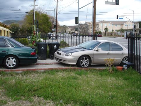 1993 Ford Taurus - Two sedans are parked on a street with trash cans and a fence in the foreground.