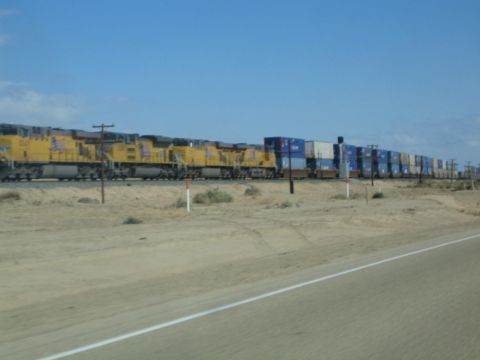 1990 Ford Bronco - long freight train with yellow locomotives and double-stacked containers runs across flat desert beside a road.