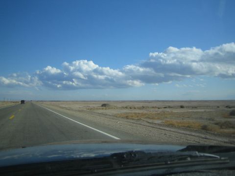 1990 Ford Bronco - straight two-lane highway runs across flat desert plains under a blue sky dotted with clouds, shot from a car.