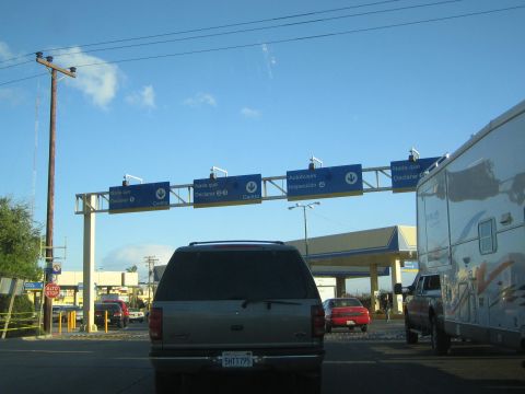 1990 Ford Bronco - Vehicles including an SUV and motorhome queue at an inspection checkpoint beneath overhead signs in Spanish.