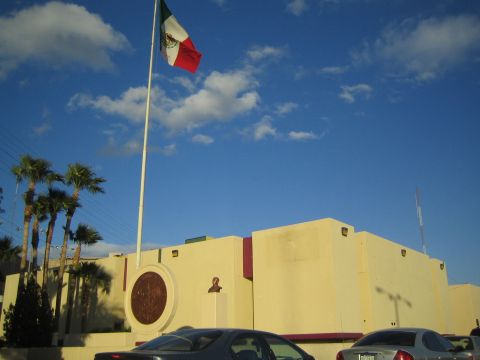 1990 Ford Bronco - large Mexican flag flies on a tall pole beside a tan government building with palm trees under a blue sky.