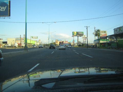 1990 Ford Bronco - wide multilane boulevard lined with billboards and shops, viewed over the dark hood of a vehicle.