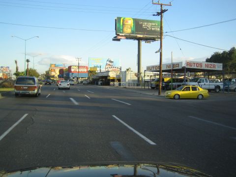 1990 Ford Bronco - yellow sedan taxi turns near the Autos Niza used car lot on a broad street with a beer billboard above.