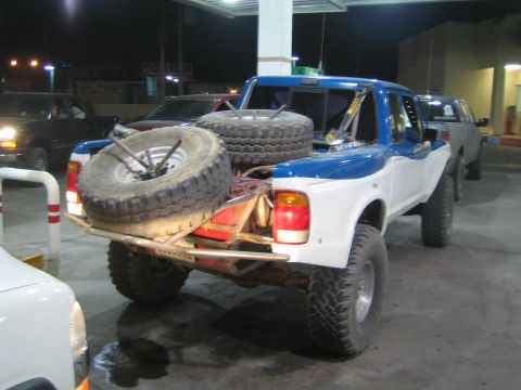1990 Ford Bronco - lifted blue-and-white prerunner pickup carrying spare off-road tires sits at a fuel station at night.