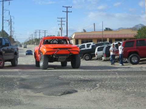 1990 Ford Bronco - orange tube-chassis Trophy Truck-style prerunner sits on a gravel lot beside a tan building as people look on.