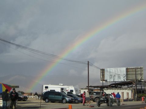 1990 Ford Bronco - bright rainbow arcs over a desert staging area with an RV, parked cars, and an off-road race truck.