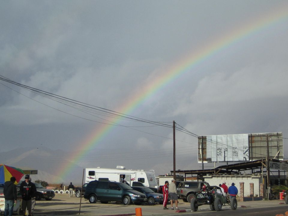 Rainbow over a desert race pit with RV, cars, and an off-road truck