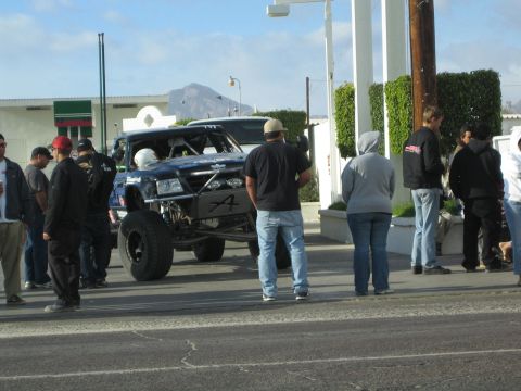 1990 Ford Bronco - black tube-chassis off-road race truck idles on a street as a crowd of spectators gathers around it.