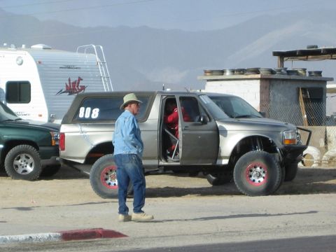 1990 Ford Bronco - man in denim and a cowboy hat stands beside a silver Ford Bronco prerunner marked 808 at a desert pit.