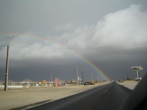 1990 Ford Bronco - rainbow arcs across a stormy sky over an open desert highway lined with sparse roadside lots.