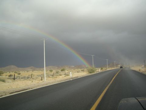 1990 Ford Bronco - vivid rainbow arcs over a two-lane desert highway lined with utility poles beneath heavy gray storm clouds.