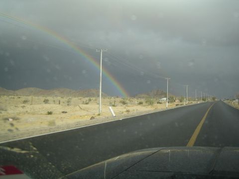 1990 Ford Bronco - rainbow descends to the desert ground beside a wet highway, viewed through a rain-flecked windshield over a dark dashboard.