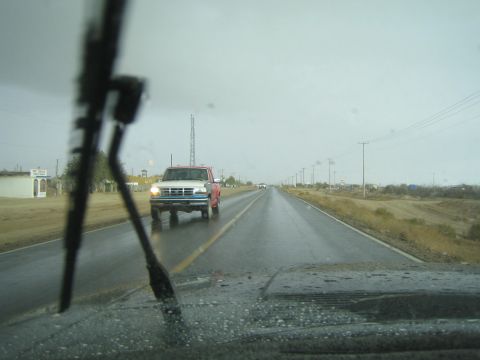 1990 Ford Bronco - blue and red pickup truck approaches on a wet two-lane road, seen through a windshield wiper from inside another vehicle.