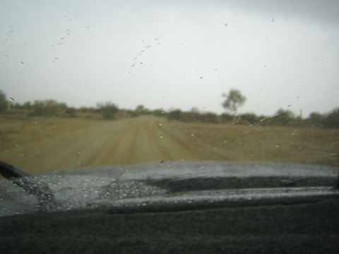 1990 Ford Bronco - faint dirt track crosses flat desert scrub under an overcast sky, viewed through a heavily rain-spotted windshield.