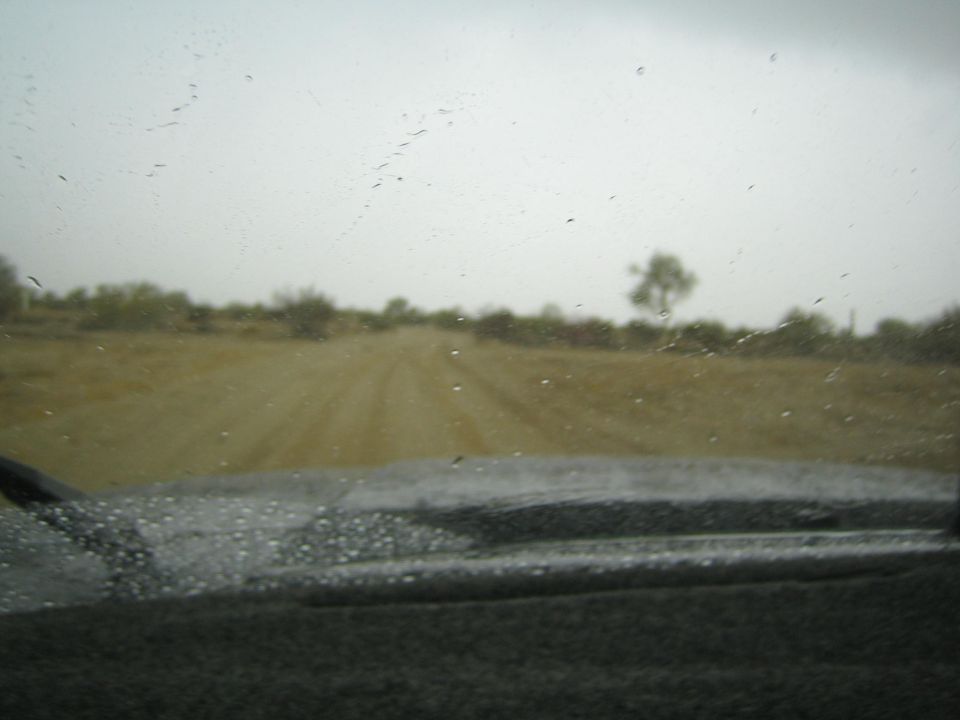Dirt road across open scrubland seen through a rain-spotted windshield and dark dashboard
