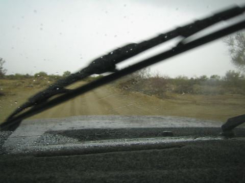 1990 Ford Bronco - sandy desert track recedes into scrub brush, seen past a raised windshield wiper and wet dashboard from inside a vehicle.