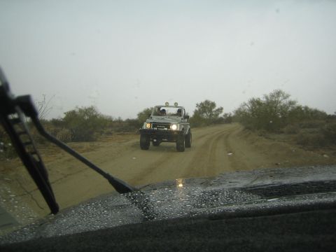 1990 Ford Bronco - white tube-frame off-road buggy with a roof light bar drives along a sandy desert track, viewed through a rain-flecked windshield.