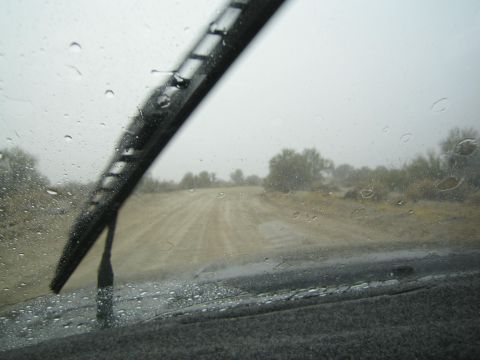 1990 Ford Bronco - dirt road stretches through desert brush, framed by a wiper blade and raindrops on the windshield of a vehicle.