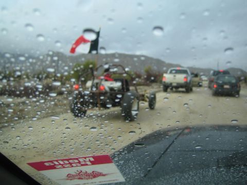 1990 Ford Bronco - Mexican flag waves over an off-road buggy and pickups parked along a sandy roadside, viewed through a rain-covered windshield with a press…