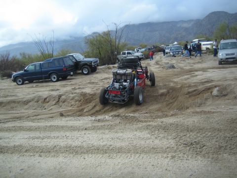 1990 Ford Bronco - red tube-frame off-road buggy sits on rough sand beside parked trucks and spectators, with cloud-covered mountains in the distance.