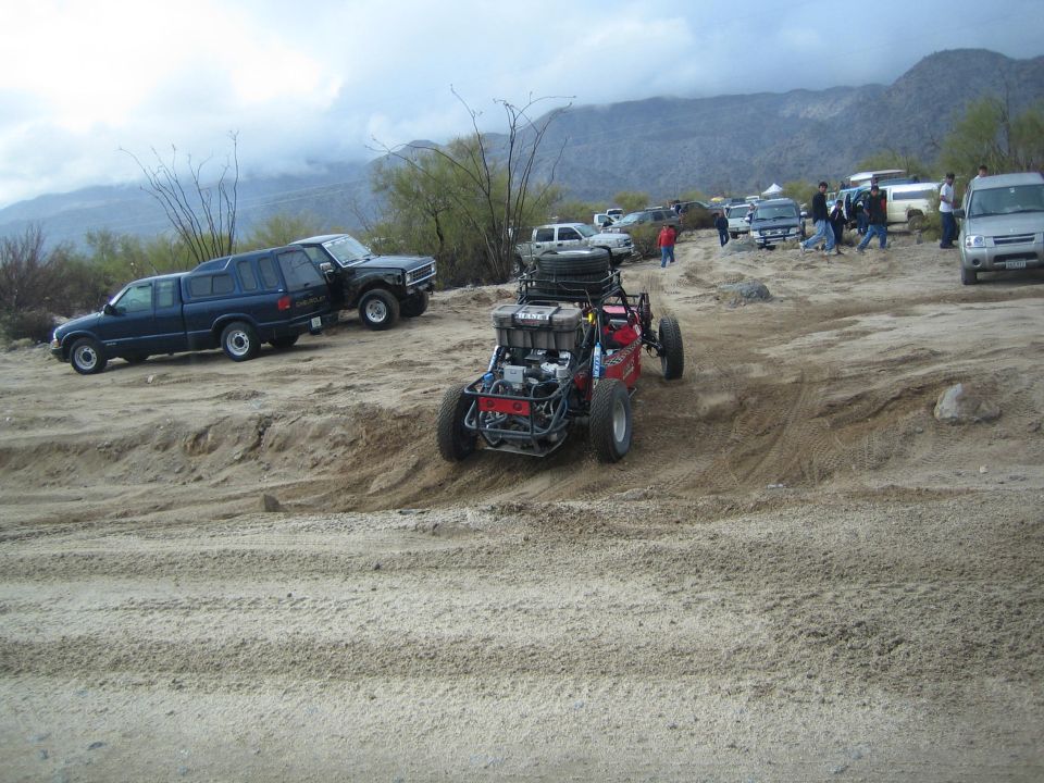 Red tube-frame off-road buggy parked on sand among trucks with mountains behind