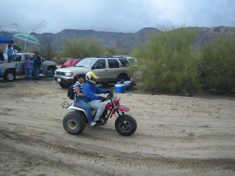 1990 Ford Bronco - Two people ride a three-wheeled ATV across sandy ground past parked SUVs and tents at a desert gathering under cloudy skies.