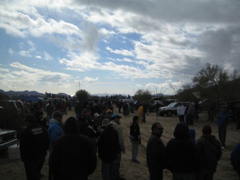 1990 Ford Bronco - dense crowd of spectators fills sandy desert terrain under towering clouds, with vehicles and pop-up canopies in the background.