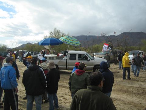 1990 Ford Bronco - Spectators in jackets gather around a silver pickup truck shaded by beach umbrellas, with a flag and cloud-shrouded mountains behind.