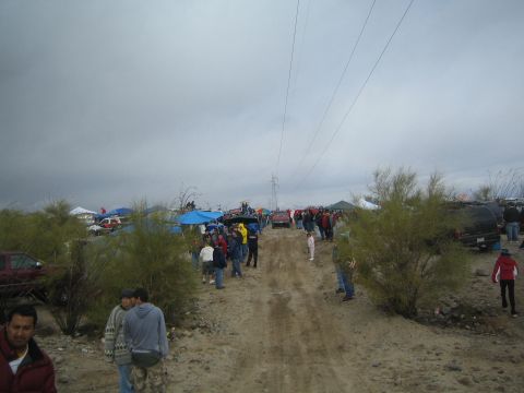 1990 Ford Bronco - Spectators walk along a sandy desert road lined with pop-up canopies and parked vehicles beneath a power transmission tower and gray sky.