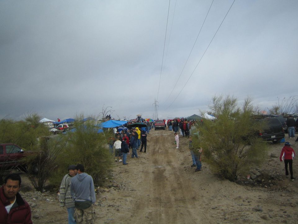 People walking a sandy desert road lined with canopies and a transmission tower