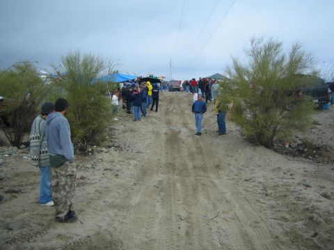 1990 Ford Bronco - People stand and walk along a wide sandy track lined with canopies and a transmission tower under an overcast desert sky.