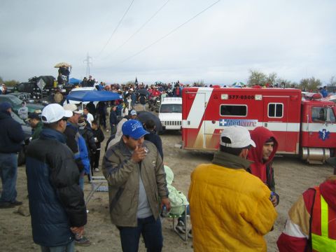1990 Ford Bronco - red Medcare ambulance is parked amid a crowd of jacketed spectators on sandy ground at an outdoor desert event.