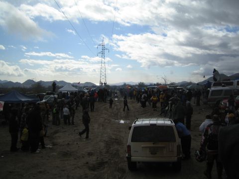 1990 Ford Bronco - Spectators and parked vehicles spread across sandy desert ground around a tall power transmission tower under a cloudy sky.
