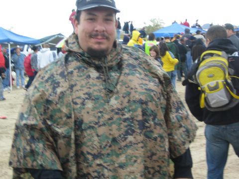 1990 Ford Bronco - smiling man wearing a digital-camo poncho and ball cap stands in the foreground of a crowded desert gathering with canopies behind.