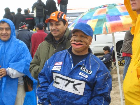 1990 Ford Bronco - man in a blue racing suit wears a comical caricature mask while standing with smiling spectators under a colorful umbrella.