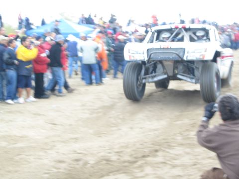 1990 Ford Bronco - white off-road race truck catches air off a sand jump as a crowd of spectators and photographers watch closely.
