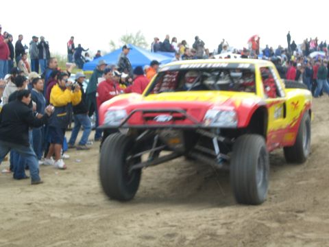 1990 Ford Bronco - red and yellow off-road race truck leaps over a sand berm as spectators and photographers line the course behind it.