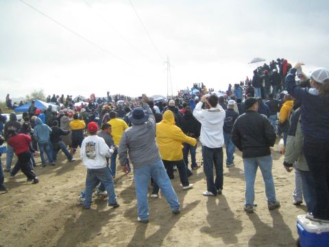 1990 Ford Bronco - large crowd in jackets watches and photographs the action across a sandy desert race course under a transmission tower and gray sky.
