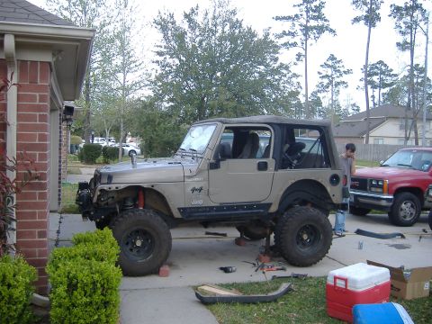2004 Jeep Wrangler - A tan 2004 Jeep Wrangler is seen in a driveway with its front end partially disassembled, next to a red GMC pickup truck.