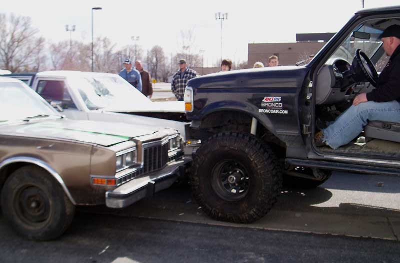Lifted Ford Bronco driving its front tire up onto a brown sedan in a parking lot crowd