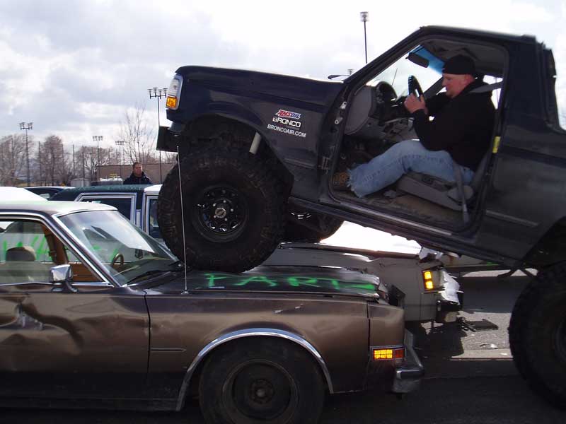 Lifted Ford Bronco front end perched on the trunk of a graffiti-tagged brown sedan, driver inside