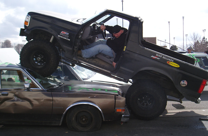 Lifted black Ford Bronco tilted up on a brown sedan in a parking lot, driver leaning out the door