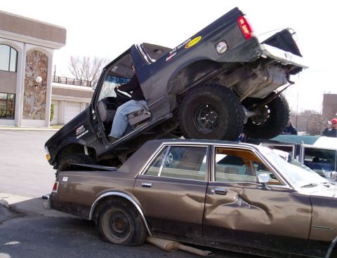 1996 Ford Bronco - A black Ford Bronco is precariously balanced on top of a severely damaged brown sedan.